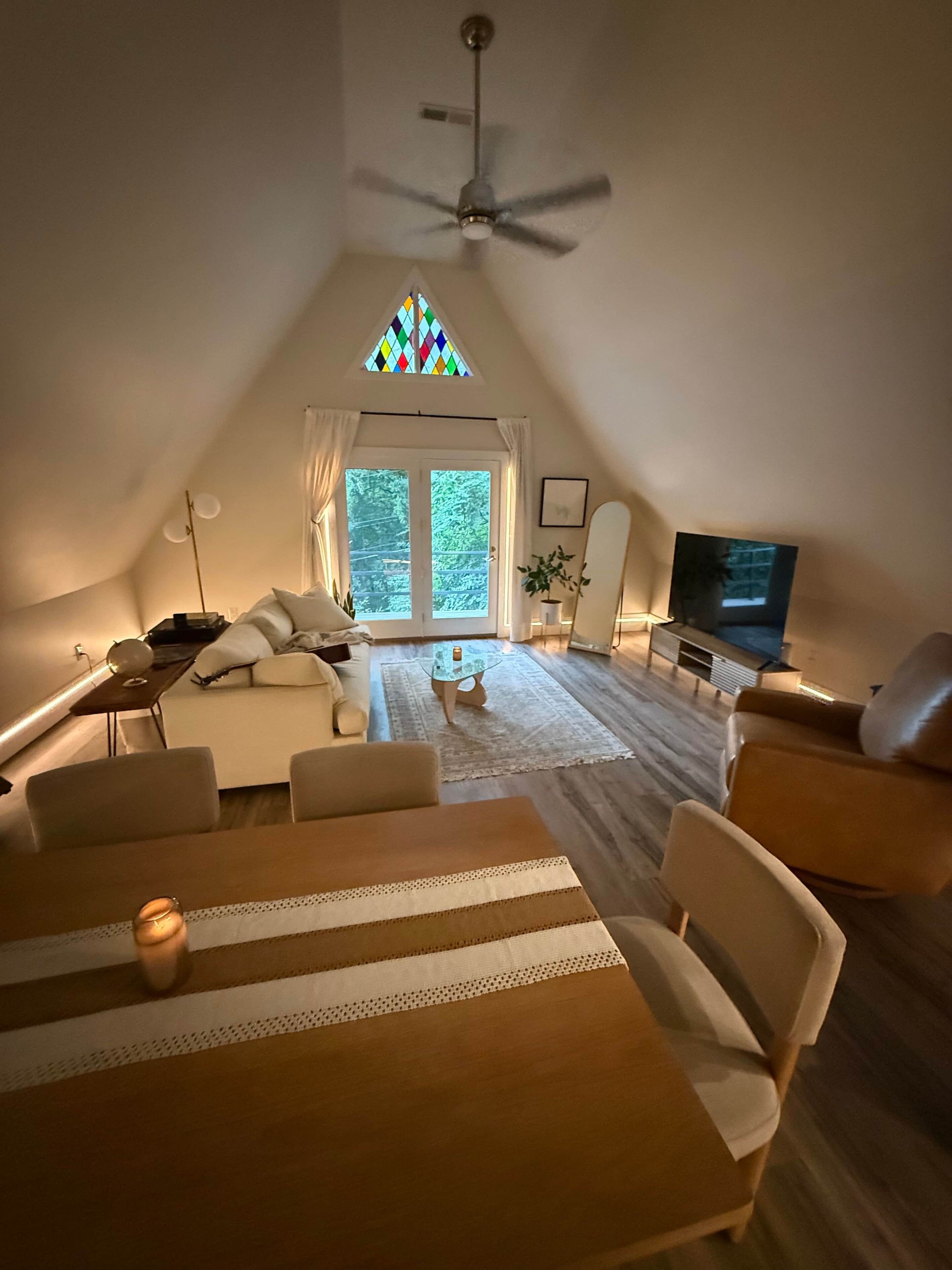 A cozy, well-lit room with a dining table, chairs, a sofa, a television, and a ceiling fan.