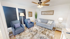 A living room with a ceiling fan, a grey sofa, a blue armchair, a patterned rug, and a wall with a 'grateful' sign. - Renovated Sandpiper Cove 2033 (Destin)
