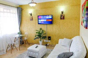 A living room with a sofa, a coffee table, a TV mounted on the wall, and a small table with chairs. - Cosy 1 bedroom Westlands -School Lane (Nairobi)