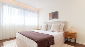 Une chambre à coucher bien rangée, avec un grand lit, une table de chevet avec une lampe et une fenêtre ornée de rideaux légers.