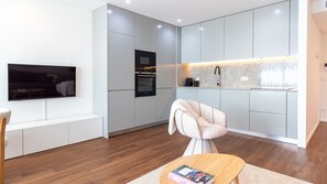 Una sala moderna con televisor de pantalla plana, un sofá, una mesita de centro con un libro, y una cocina con microondas y hervidor.