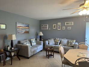 A living room with two sofas, a coffee table, a dining table with chairs, a ceiling fan, and wall-mounted artwork. - Wonderful first floor 2-bedroom condo with AC in beautiful Marco Island (Marco Island)