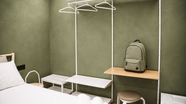 Una camera d'albergo con un letto, un comodino, un appendiabiti, una panca, uno sgabello e uno scaffale con uno zaino.