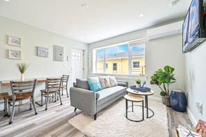 Ein modernes Wohnzimmer mit einem grauen Sofa, Holzstühlen, einem kleinen runden Tisch und einem großen Fenster mit Blick auf Gebäude.