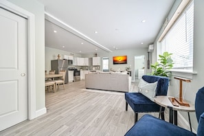 A modern kitchen with white cabinets, a dining area with a table and chairs, a living room with a sofa and a TV, and a large window with blinds. - Seaside Serenity: Salty Shore Retreat (Wildwood)