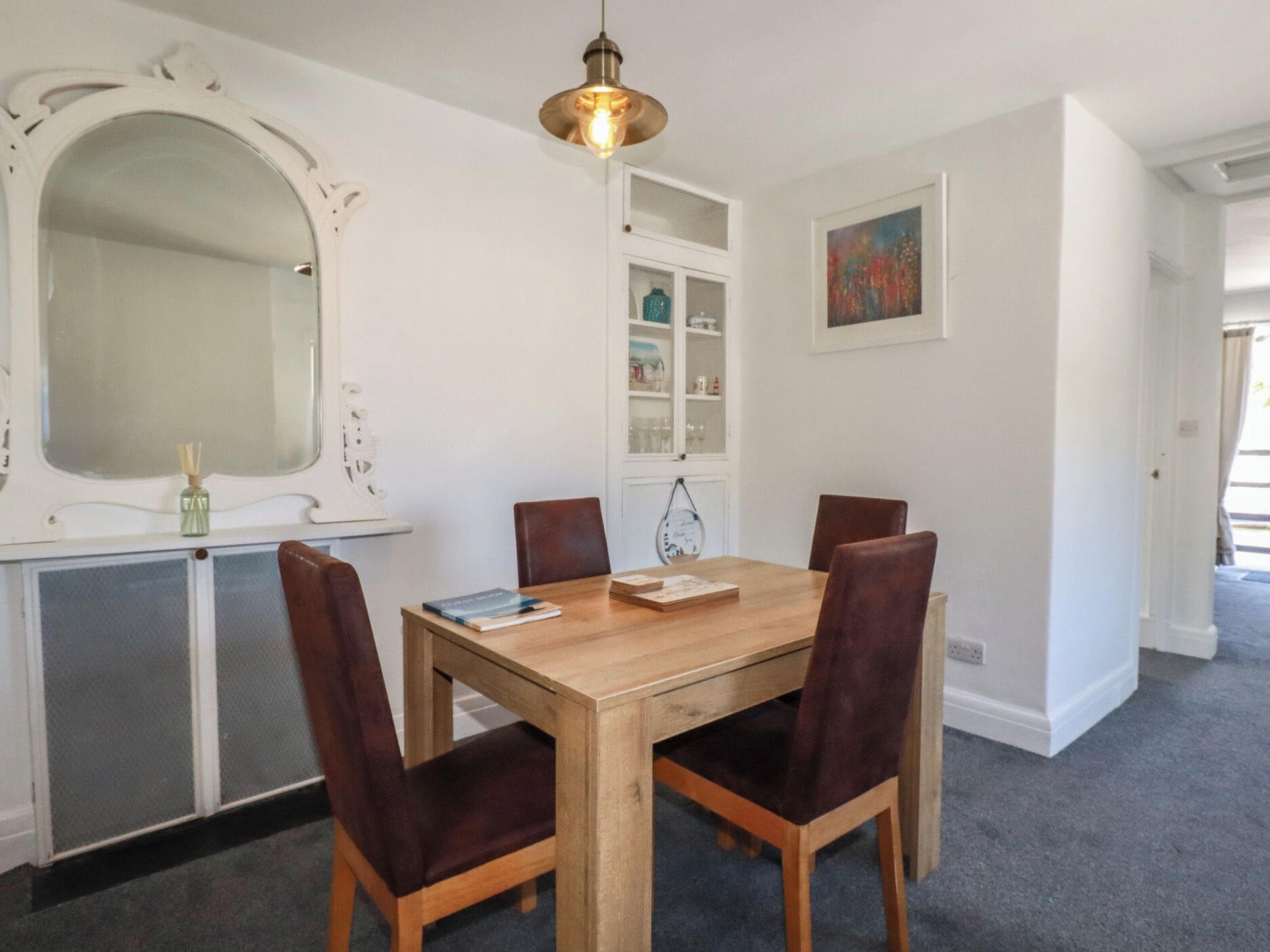 Un coin repas avec une table en bois et quatre chaises, un luminaire suspendu, un grand miroir et un tableau encadré accroché au mur.