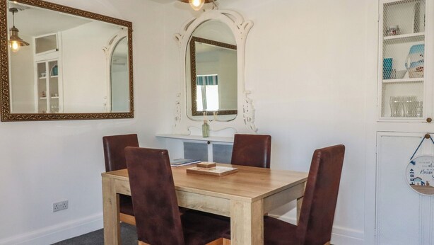 A dining room with a wooden table and four chairs, a mirror, and a shelf with glassware. - Harbourside Retreat (Looe)