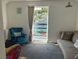 Quarto com poltrona azul, sofá, vista para um porto com barcos e janela com cortinas.