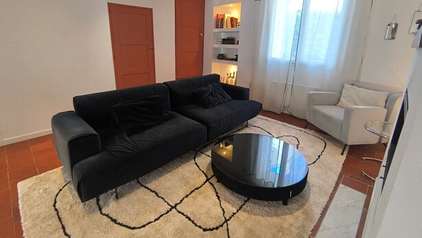 A living room with a black sofa, a white armchair, a glass coffee table, and a patterned rug. - Fajureya House (Vaumeilh)