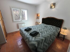 Ein Schlafzimmer mit einem Bett, einem Fenster, einer Heizung, einem Nachttisch mit Lampe und einem an der Wand befestigten Regal.