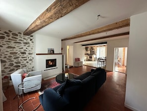 A living room with a fireplace, stone wall, and a view into the kitchen. - Fajureya House (Vaumeilh)