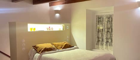 A bedroom with a bed, wooden ceiling beams, a window with curtains, and a wall-mounted light fixture.