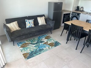 Ein kompakter Wohnraum mit einer Couch, einem Esstisch mit Stühlen, einer Küchenzeile mit Kühlschrank und einer Waschmaschine.