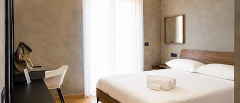 A hotel room with a bed, a desk, a chair, a hanging coat rack, and a large window with curtains.