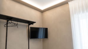 Una habitación con televisor de pantalla plana en la pared, un perchero metálico negro y una ventana con cortinas traslúcidas.