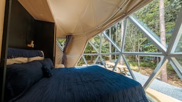 A tented accommodation with a bed, a table, and a view of the forest.