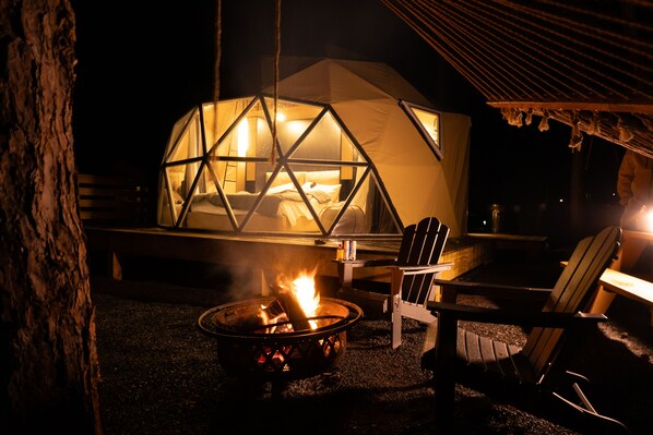A geodesic dome structure with a fire pit in the foreground, a chair, and a table.