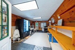 Quarto com paredes de madeira, uma geladeira preta, uma mesa com cadeiras, um tapete azul no chão e uma janela com persianas.
