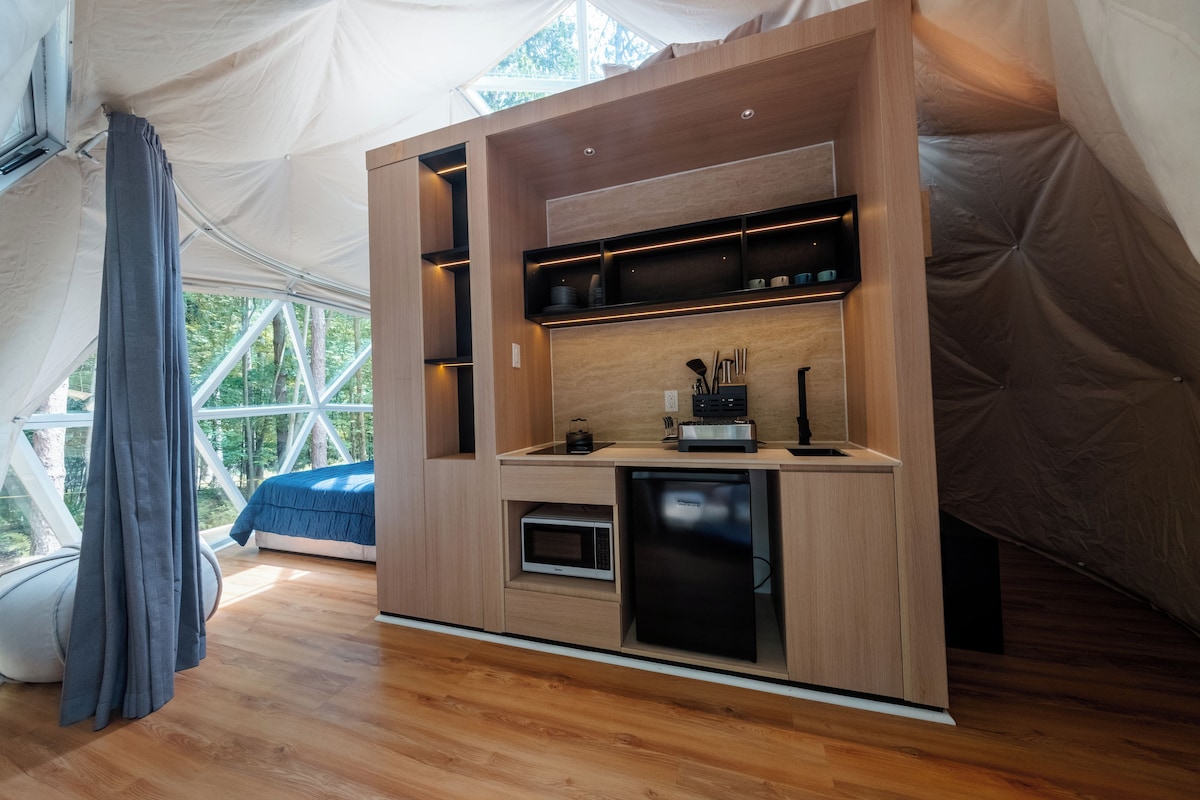 Una cocina compacta con electrodomésticos integrados y un refrigerador pequeño, ubicada en una estructura tipo tienda con vistas a árboles a través de amplios ventanales.
