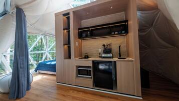 Una cocina compacta con electrodomésticos integrados y un refrigerador pequeño, ubicada en una estructura tipo tienda con vistas a árboles a través de amplios ventanales.