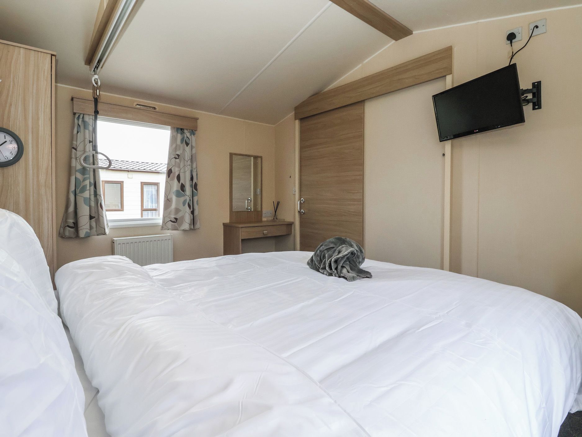 A bedroom with a bed, a television mounted on the wall, a window with curtains, and a clock on the wall.