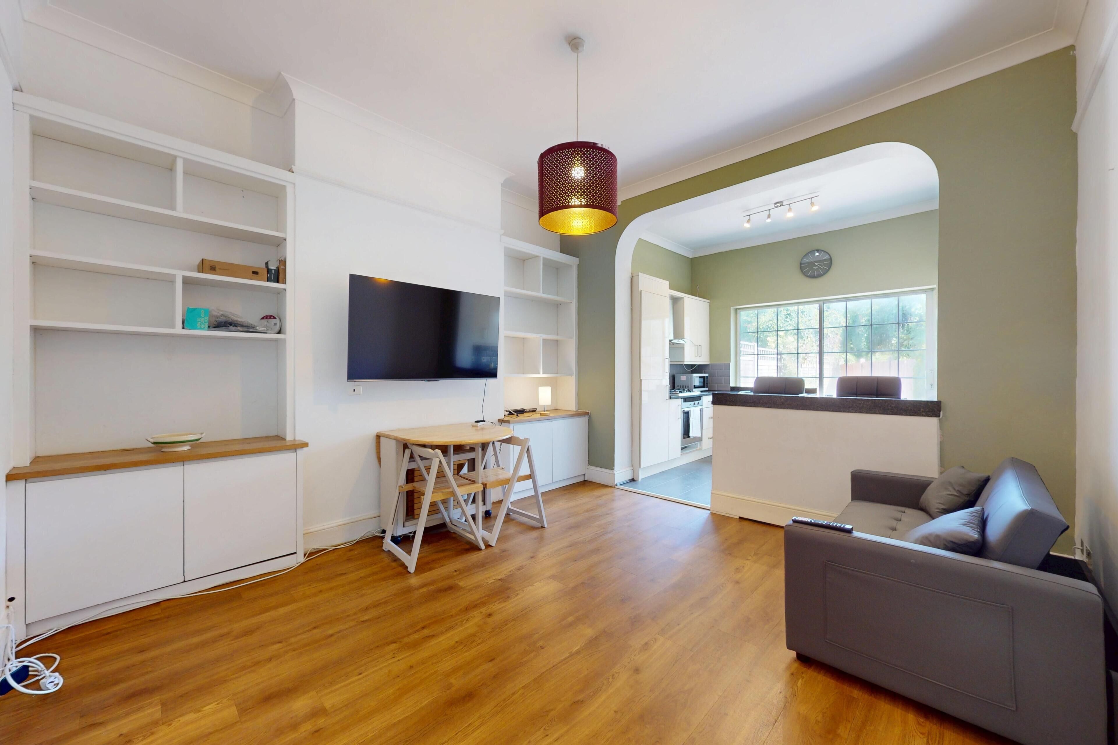 A modern living room with a flat-screen TV, wooden flooring, a grey sofa, and a dining area with a table and chairs.