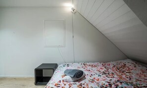 Ein kleines Zimmer mit einem Einzelbett, schrägem Dach, Nachttisch und einer Wandleuchte.