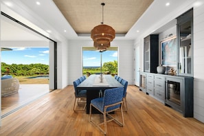 A modern dining area with a large table, blue upholstered chairs, and a view of a lush landscape through large windows. - Makani Kai by Gather: Brand-New Luxury Home w/ Private Pool & Spa, AC, Chef’ (Koloa)