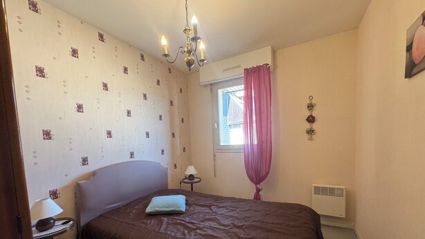 Makuuhuoneessa on sänky, yöpöytä, lamppu, ikkuna, jossa on verhot, ja seinään asennettu ilmanvaihtoaukko.