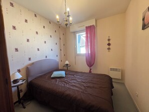 Ein Schlafzimmer mit einem Bett, einem Nachttisch, einer Lampe, einem Fenster mit Vorhängen und einem Wandlüfter.