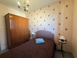 A bedroom with a bed, a wardrobe, a bedside table with a lamp, and patterned wallpaper. - Apartment in the heart of Ouistreham beach (Ouistreham)