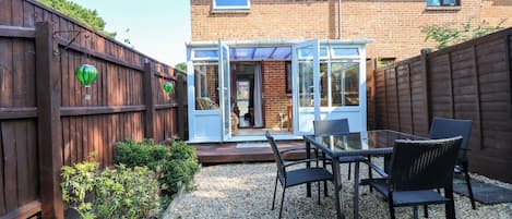 A patio with a glass table and chairs, a wooden fence, and a brick house in the background.