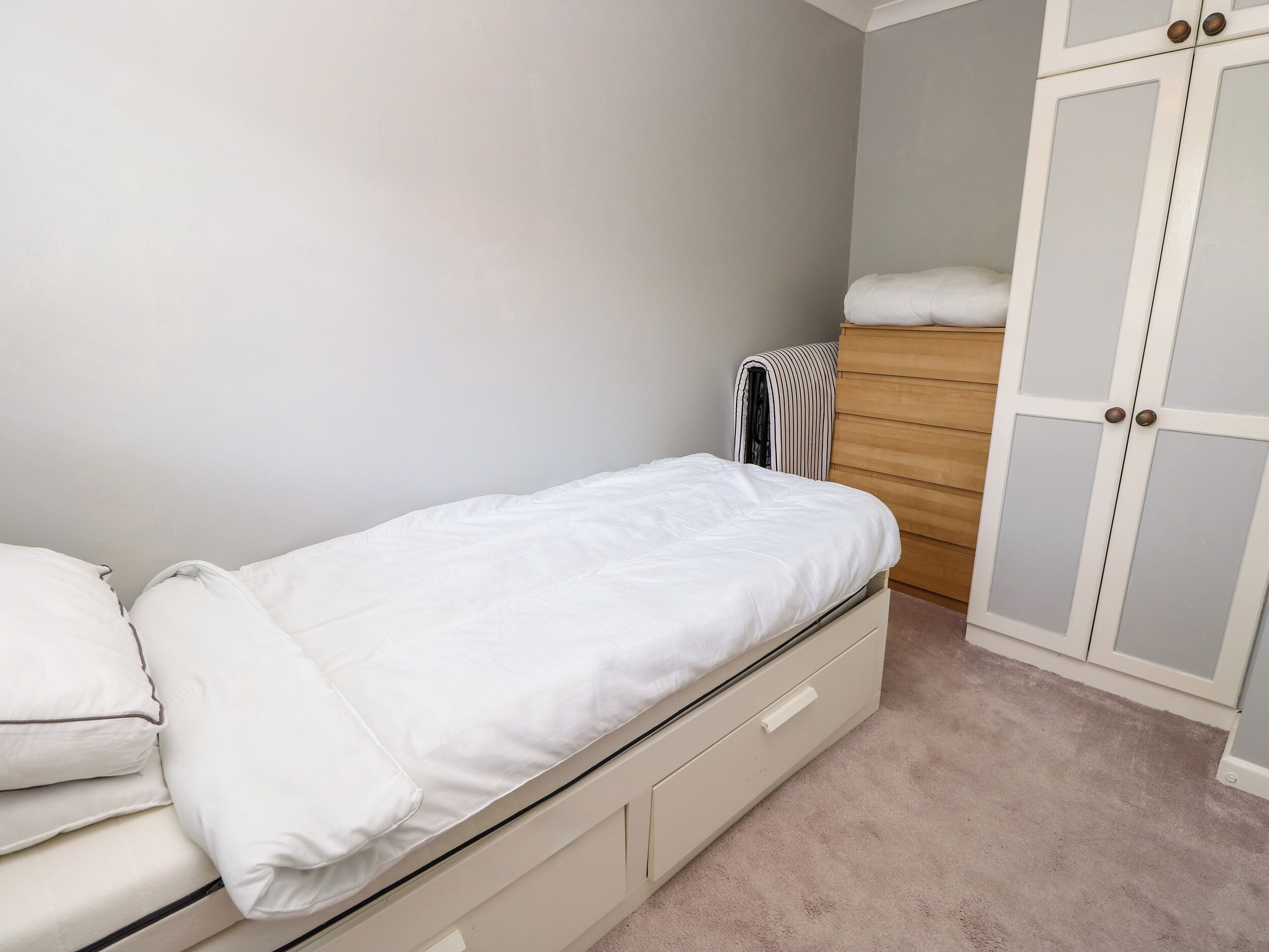 A single bed with a white mattress and pillows, a wooden dresser, and a wardrobe with frosted glass panels.