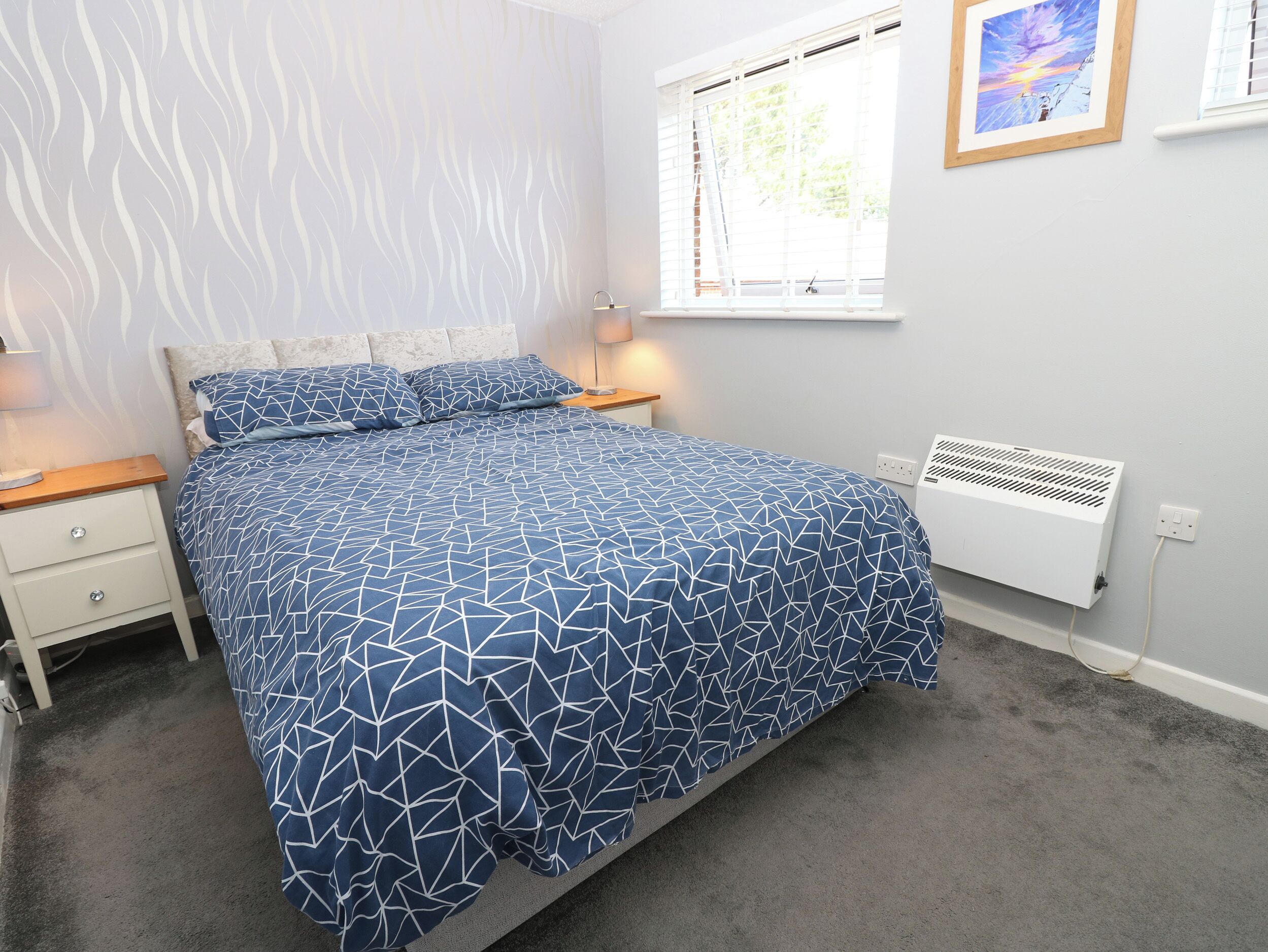 A bedroom with a bed, bedside tables, a window, a radiator, and a wall with a painting.