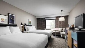 Quarto de hotel com duas camas, uma escrivaninha, televisão e janela com persianas.