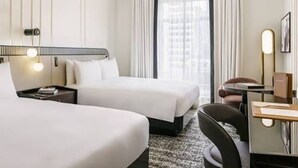 Quarto de hotel com duas camas, uma escrivaninha, uma cadeira e uma janela grande com cortinas.