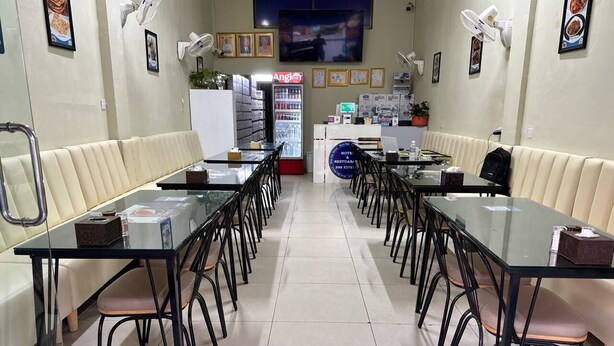 Uma sala de jantar de hotel com mesas de vidro e cadeiras pretas, um refrigerador e ventiladores fixados na parede.