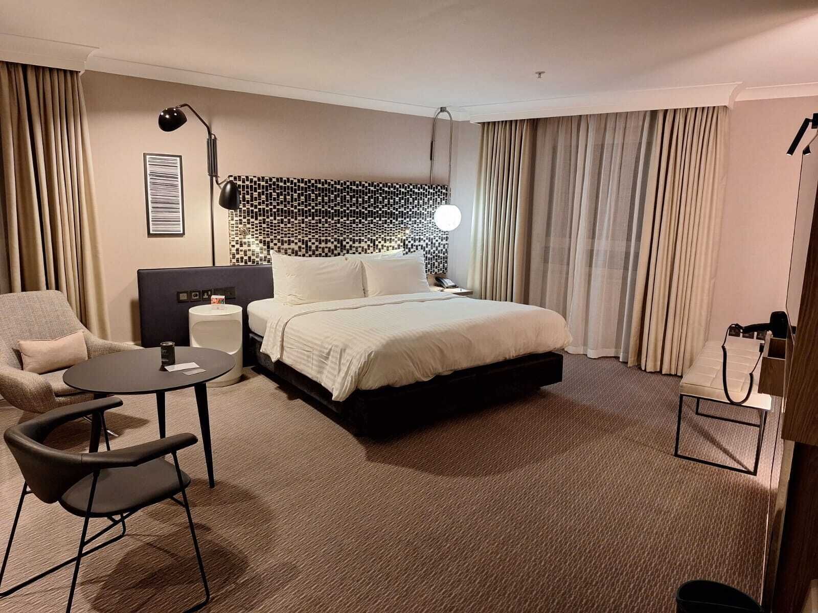 A hotel room with a bed, a desk, a chair, a lamp, and a window with curtains.