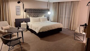 Quarto de hotel com cama, escrivaninha, cadeira, abajur e janela com cortinas.
