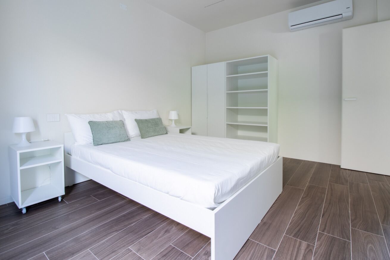 A bedroom with a white bed, a nightstand, a wardrobe, and an air conditioning unit.