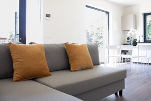 Ein modernes Wohnzimmer mit einem grauen Ecksofa, zwei senffarbenen Kissen, einem weißen Essbereich mit Tisch und Stühlen sowie einem großen Fenster, das natürliches Licht hereinlässt.