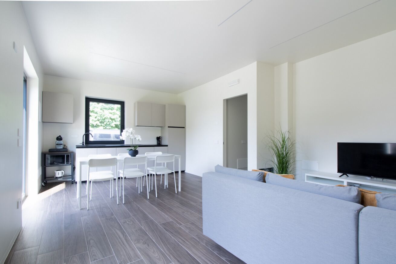 Ein modernes Wohnzimmer mit grauem Sofa, einem Flachbildfernseher und einem Essbereich mit weißen Stühlen und einem Tisch.