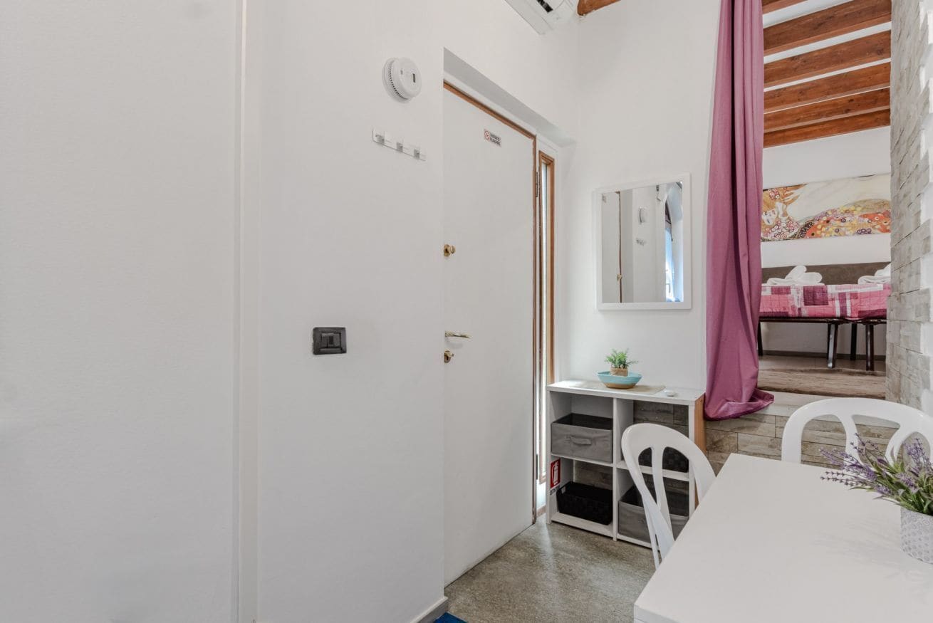 Ein Zimmer mit weißer Tür, einem kleinen Schreibtisch mit Spiegel, einem rosa Vorhang und einem Tisch mit Stühlen.
