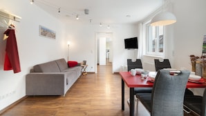 A modern living room with a grey sofa, a dining table with chairs, a flat-screen TV mounted on the wall, and a window with curtains. - Gaiser Apartment, Urach Waterfall, Swabian Alb (Bad Urach)