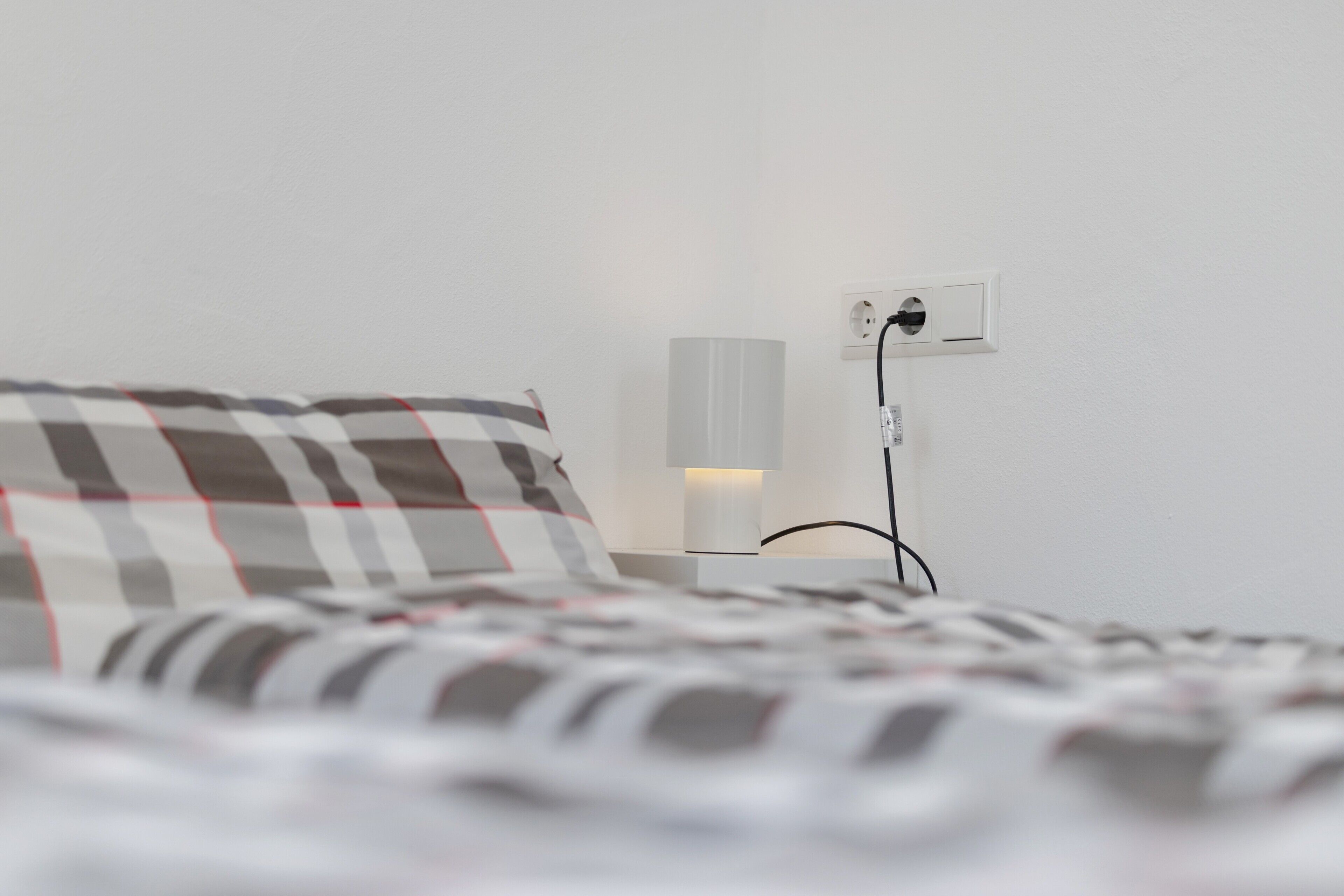 Una cama con una colcha a cuadros y una mesita de noche con una lámpara y un enchufe.