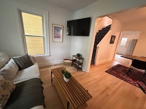 A living room with a sofa, a coffee table, a TV mounted on the wall, a staircase leading to another room, and a dining area with a table and chairs. - Cozy 3-bedroom house in the heart of the east side of Madison (Madison)