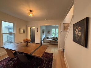 Ein Essbereich mit einem Holztisch, Stühlen und einer Vase mit Trockenblumen. Ein Flur führt in ein Wohnzimmer mit einer Couch und einem Fenster.