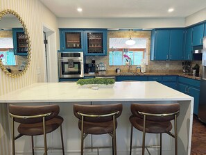 A modern kitchen with blue cabinets, a white island, and three bar stools. - Be A Nomad | The Seabreeze 5BR Pool House (Jacksonville Beach)