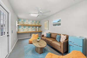 A compact living space with a brown leather sofa, a round wooden coffee table, a blue refrigerator, and a ceiling fan. - Be A Nomad | The Seabreeze 5BR Pool House (Jacksonville Beach)