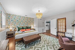 Ein Schlafzimmer mit einem Bett, Nachttischen, einer Bank, einem Sessel, einer Hängeleuchte und einem gemusterten Teppich.